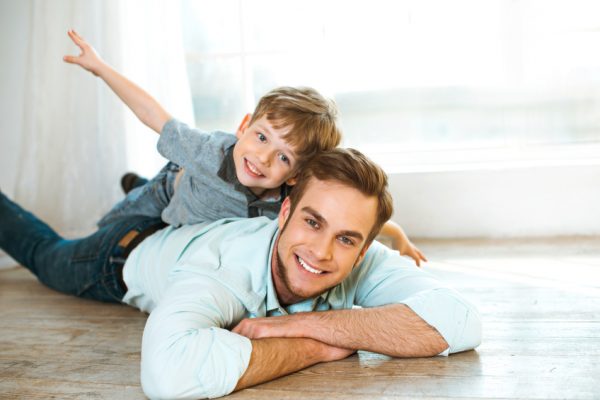 Nice family photo of little boy and his father. Boy and dad smiling and lying on wooden floor. Boy riding piggyback